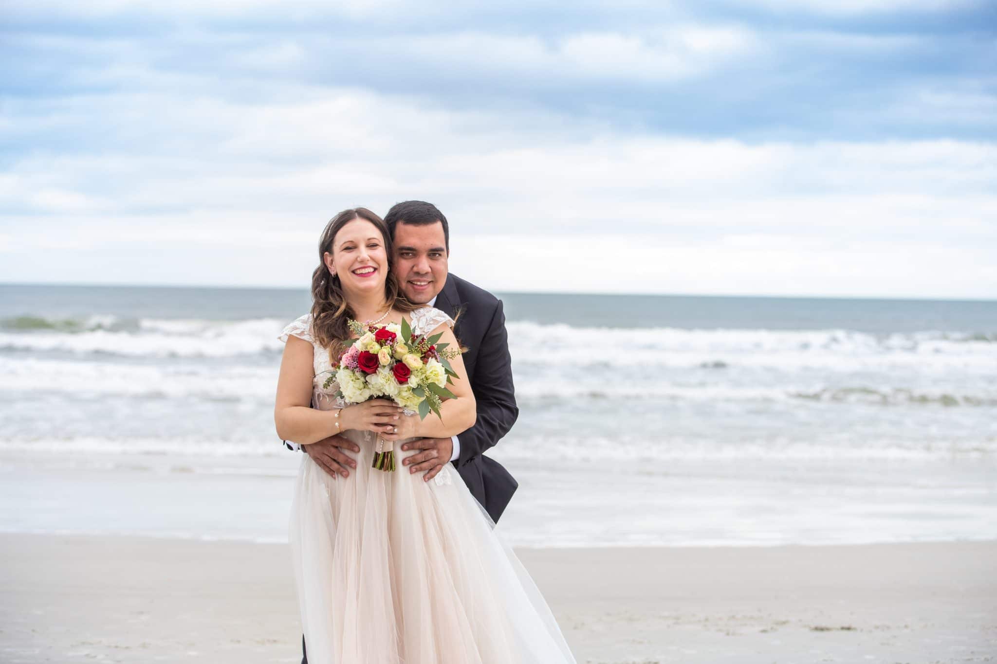 beach wedding photography of a young couple in daytona beach with a wedding dress, bouquet. Groom is standing behind the bride embracing her as they pose for a photo on the beach